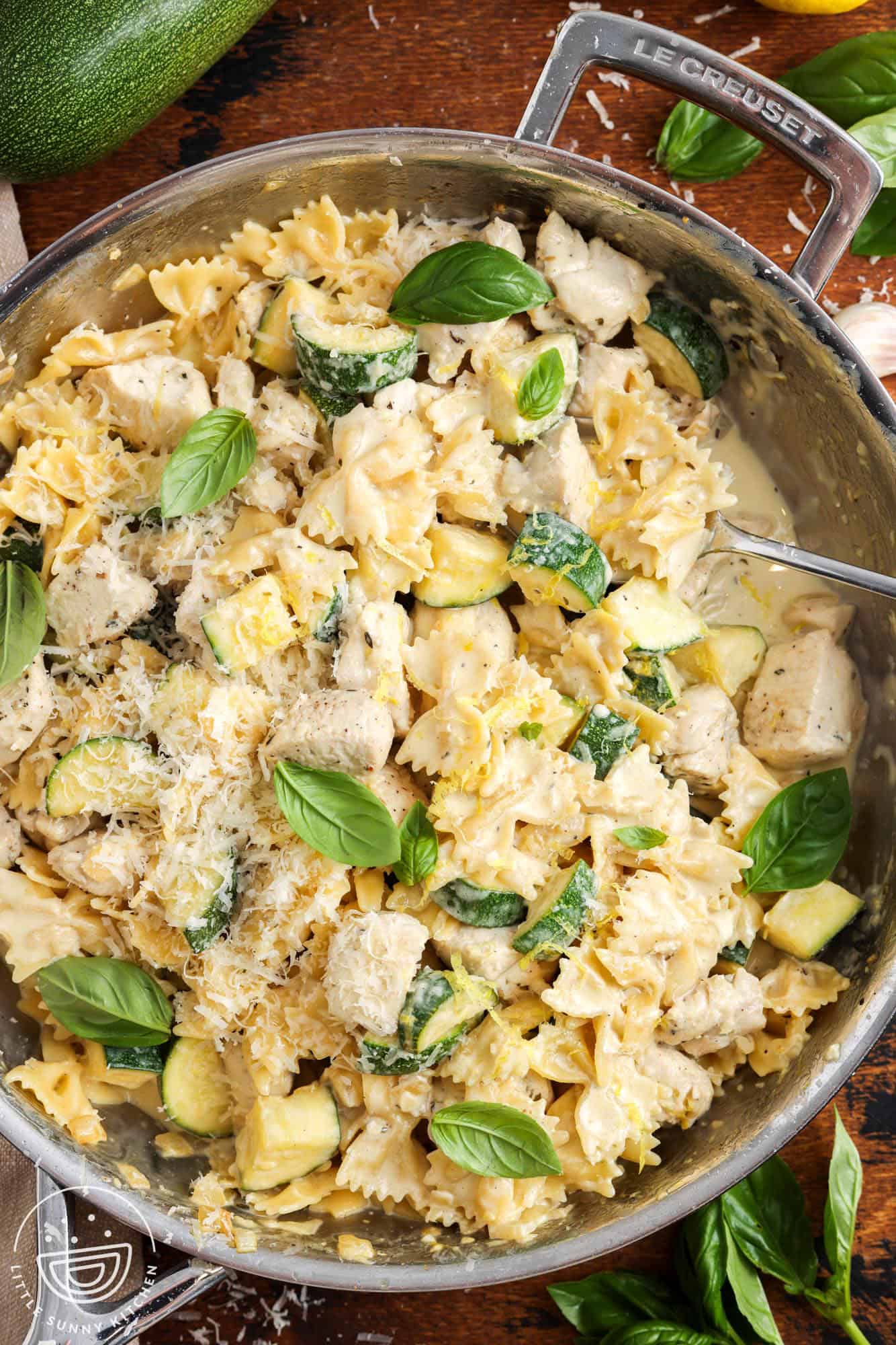 creamy chicken and zucchini pasta served from a stainless steel skillet with a spoon. The pasta is garnished with whole basil leaves and grated parmesan.