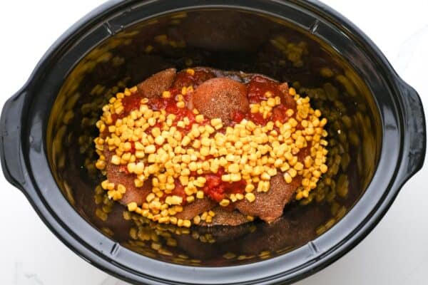 chicken topped with seasonings, salsa, and corn, in a black crockpot.