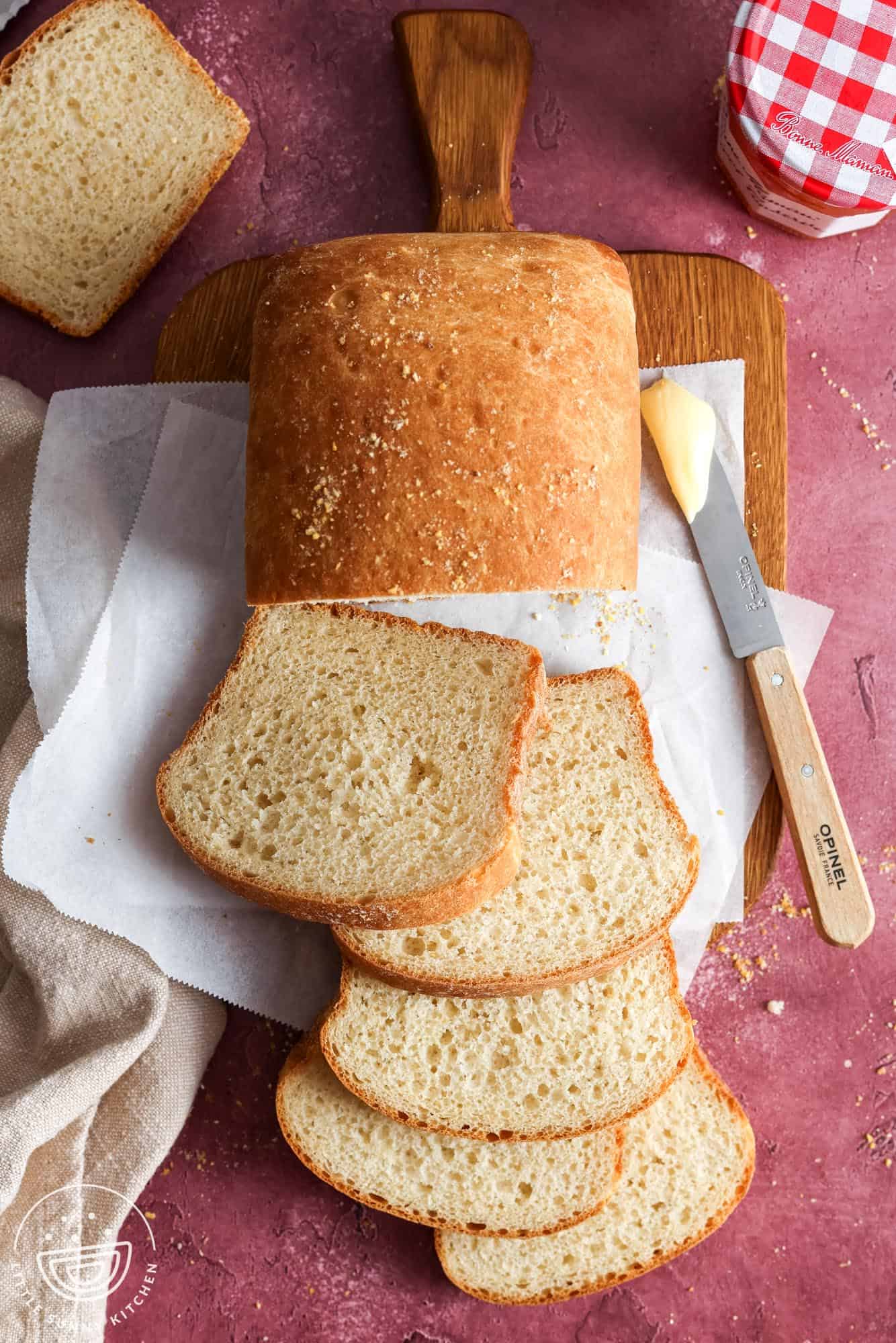 overhead view of a loaf of English Muffin Bread sliced on a wooden cutting board. With the bread is a table knife holding butter and a jar of jam.