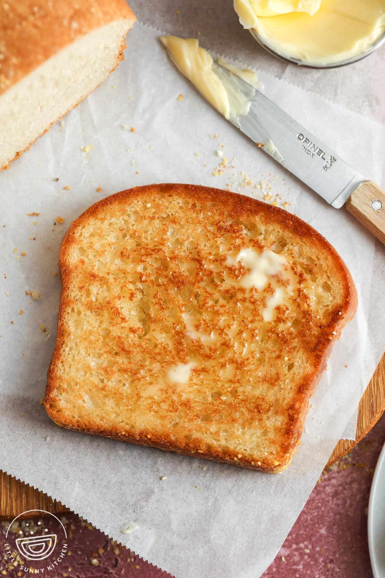 a toasted slice of english muffin style bread on a piece of parchment paper. A knife has just spread butter on the toast and the butter is is melted.