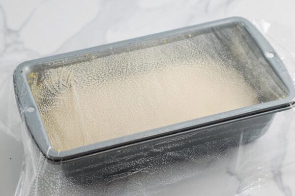 dough rising in a metal loaf pan, covered with greased plastic wrap.