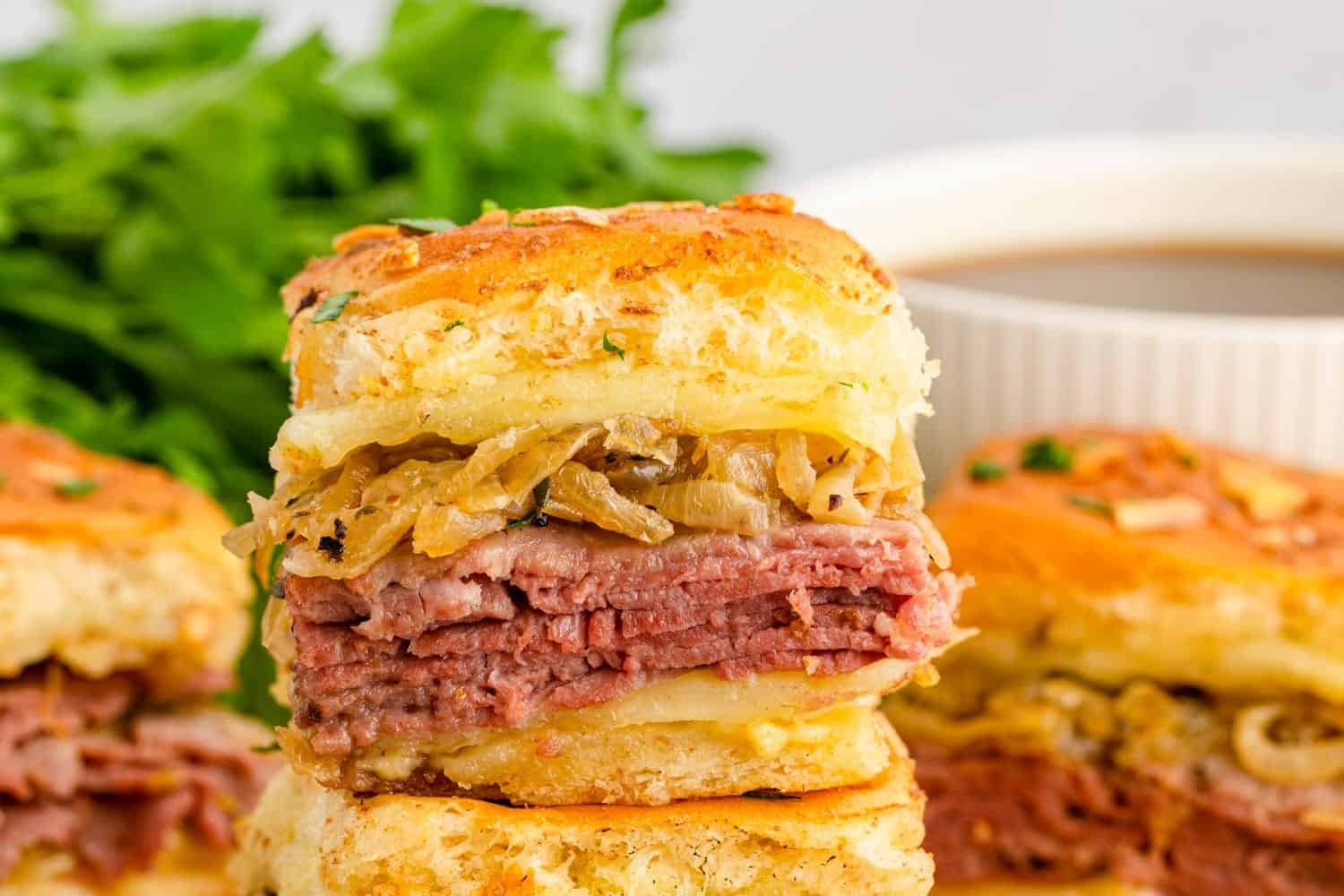 Close-up of a French dip slider with layers of roast beef, melted cheese, and caramelized onions on a golden toasted bun, with au jus dipping sauce in the background.