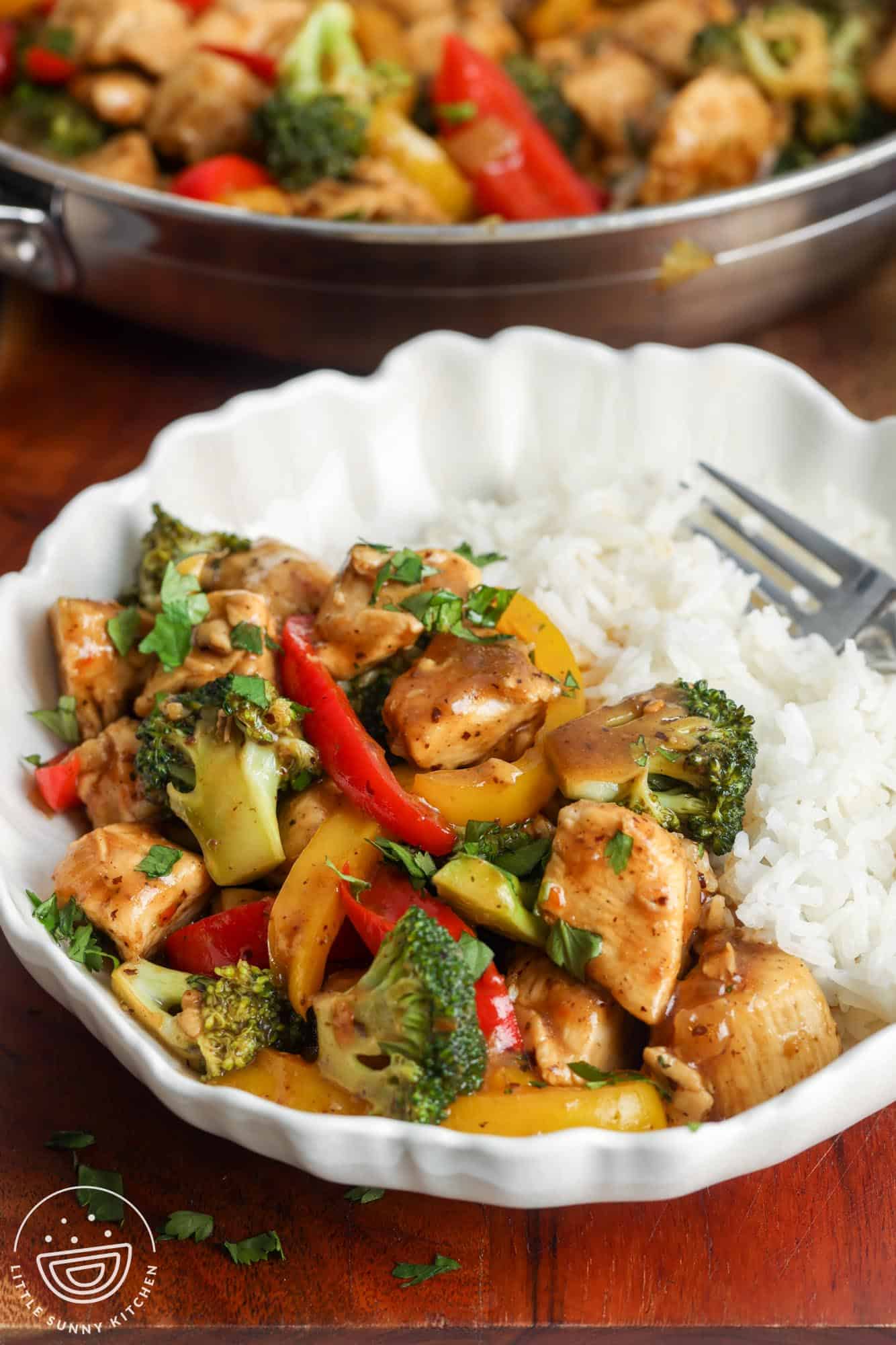 a white ceramic bowl of half rice and half hoisin chicken stir fry. In the background is a skillet of chicken with veggies.