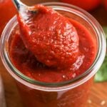 A spoonful of thick homemade pizza sauce being lifted from a glass jar, showing its rich red color and smooth texture.