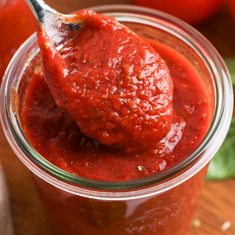 A spoonful of thick homemade pizza sauce being lifted from a glass jar, showing its rich red color and smooth texture.