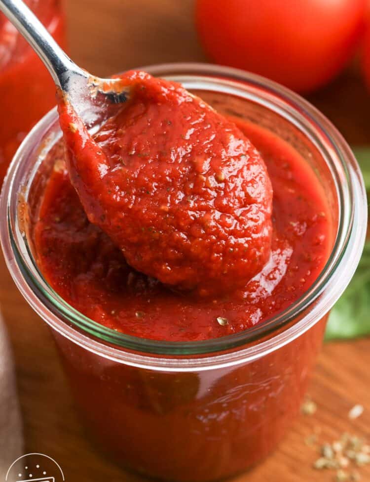 A spoonful of thick homemade pizza sauce being lifted from a glass jar, showing its rich red color and smooth texture.