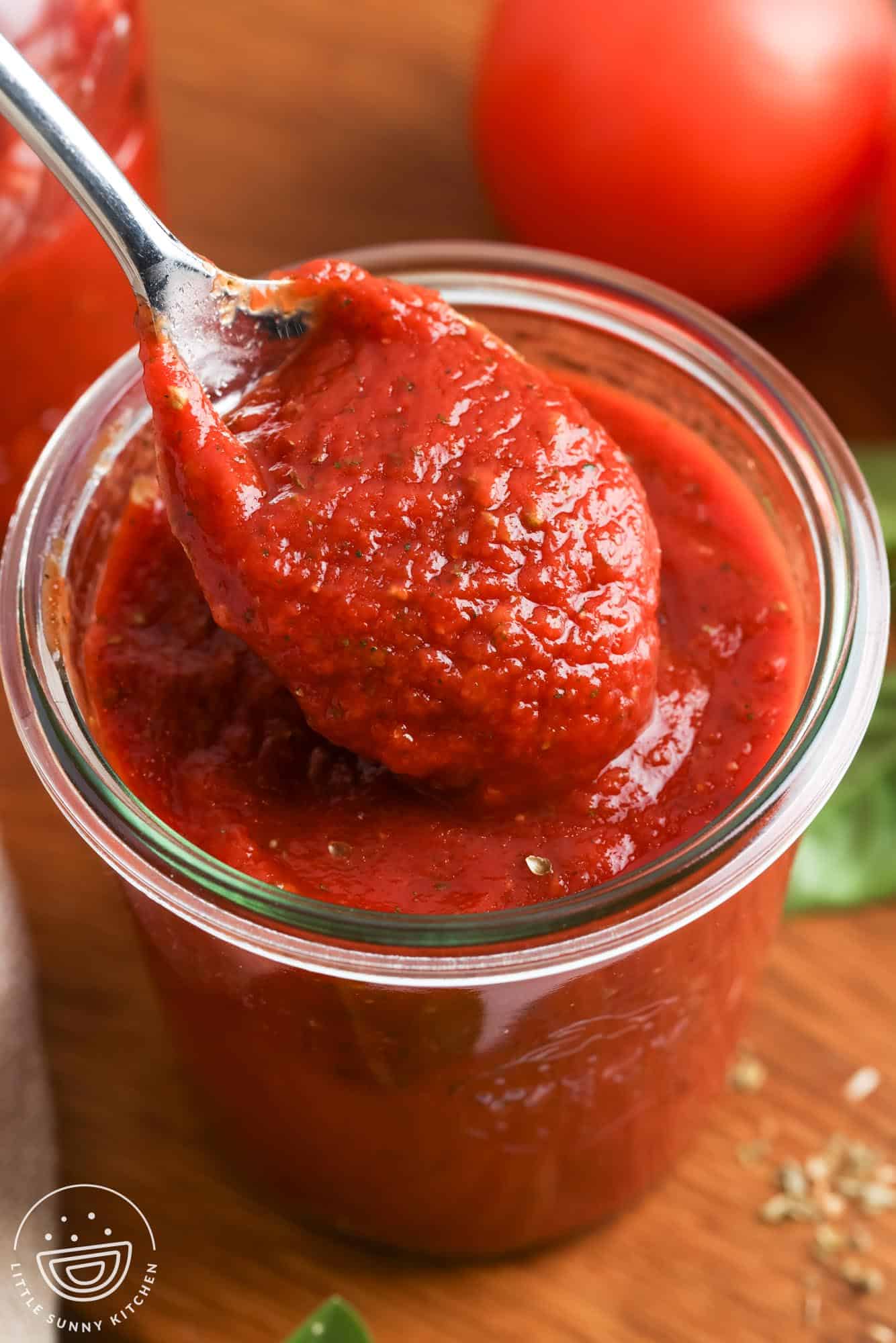 A spoonful of thick homemade pizza sauce being lifted from a glass jar, showing its rich red color and smooth texture.
