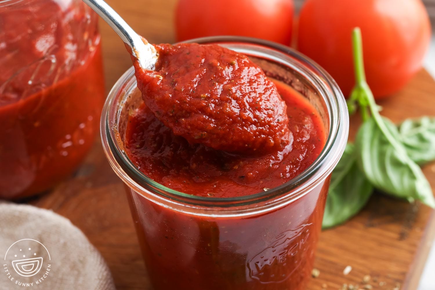 A spoonful of pizza sauce resting over a glass jar with tomatoes and fresh basil in the background.