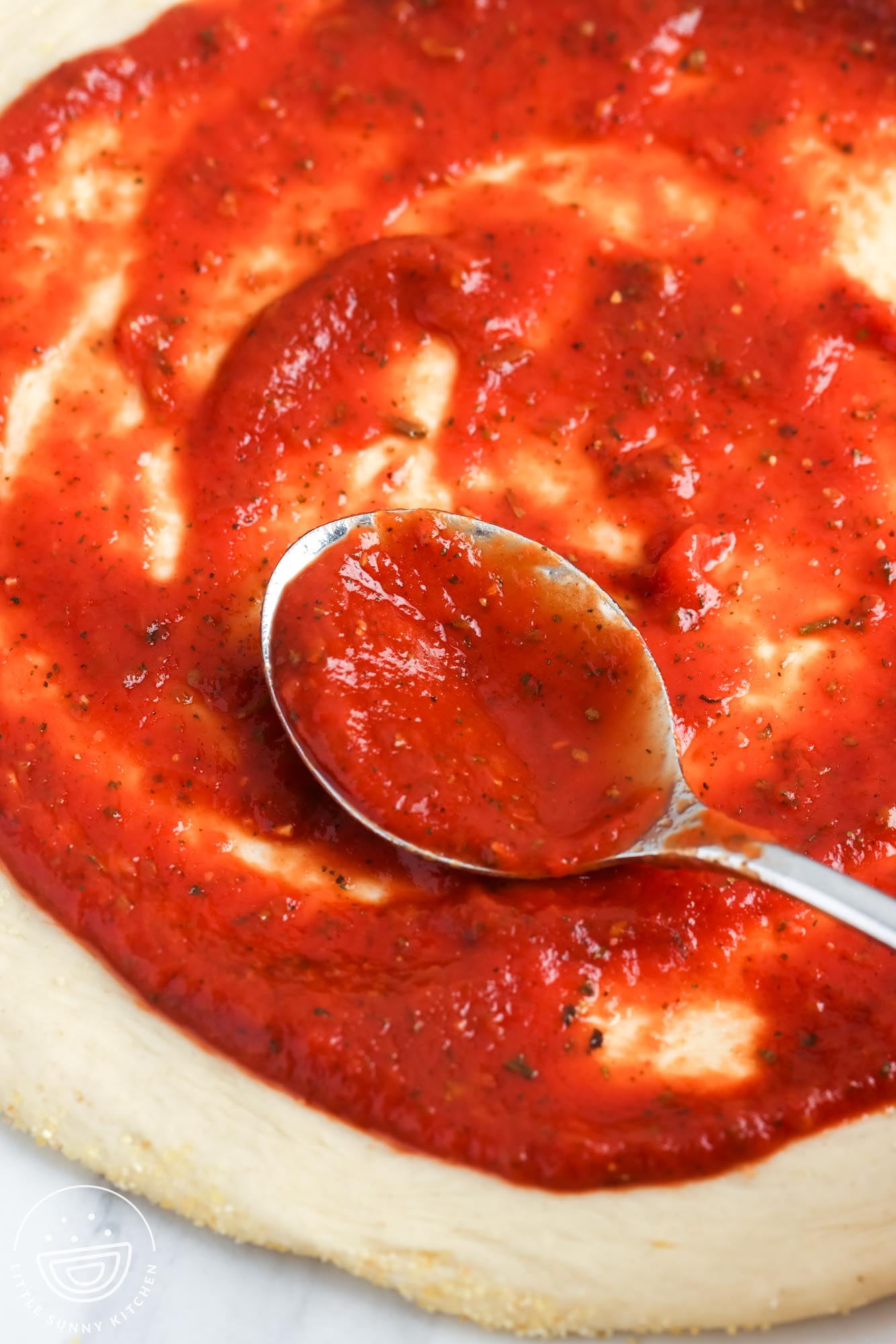 A spoon spreading bright red homemade pizza sauce over raw pizza dough.