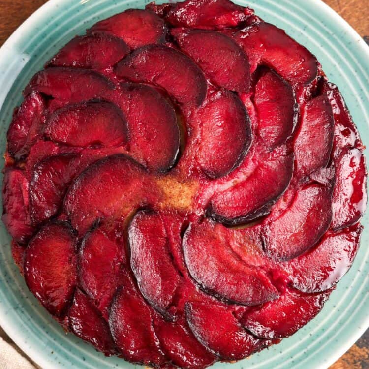 Overhead view of a whole spiced plum upside down cake on a teal plate, topped with a spiral pattern of glossy red plum slices.