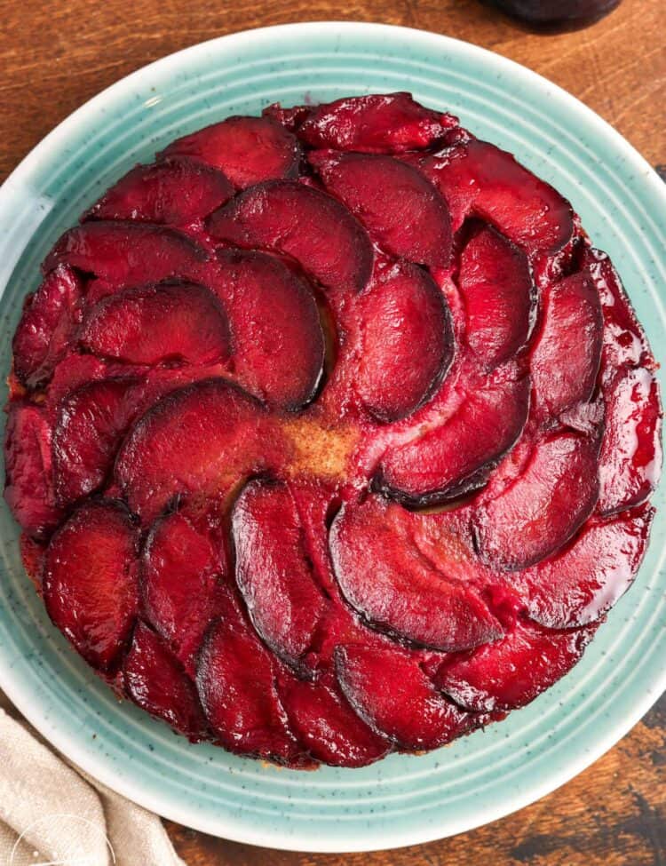 Overhead view of a whole spiced plum upside down cake on a teal plate, topped with a spiral pattern of glossy red plum slices.