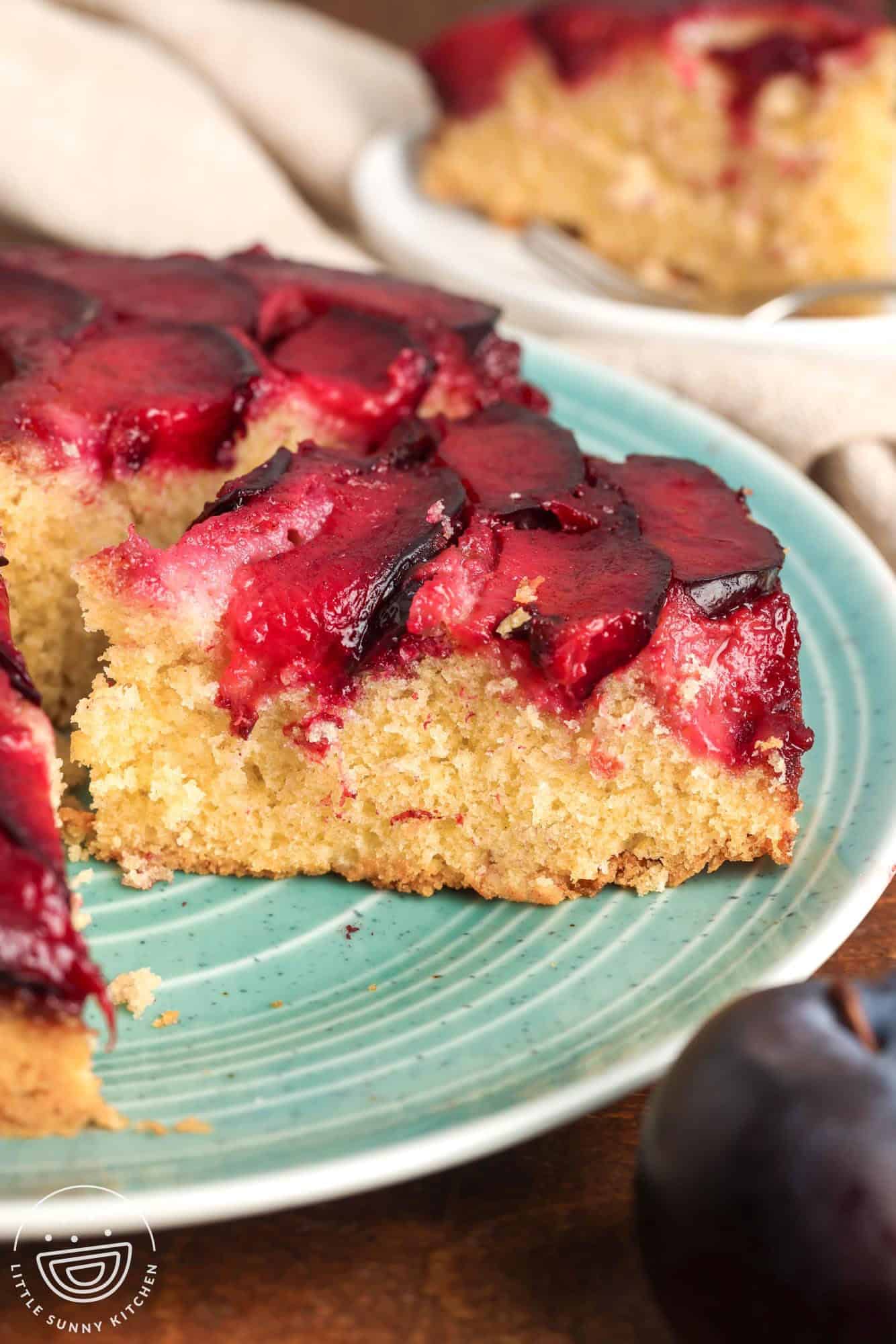 Side view of a slice of spiced plum upside down cake on a teal plate, showing the fluffy golden crumb and a topping of caramelized red plums.