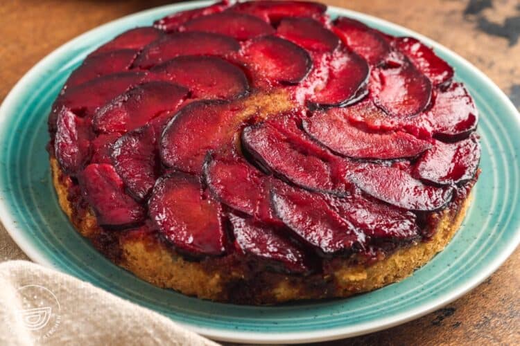 Whole spiced plum upside down cake on a teal plate, topped with glossy caramelized plum slices arranged in a spiral pattern.