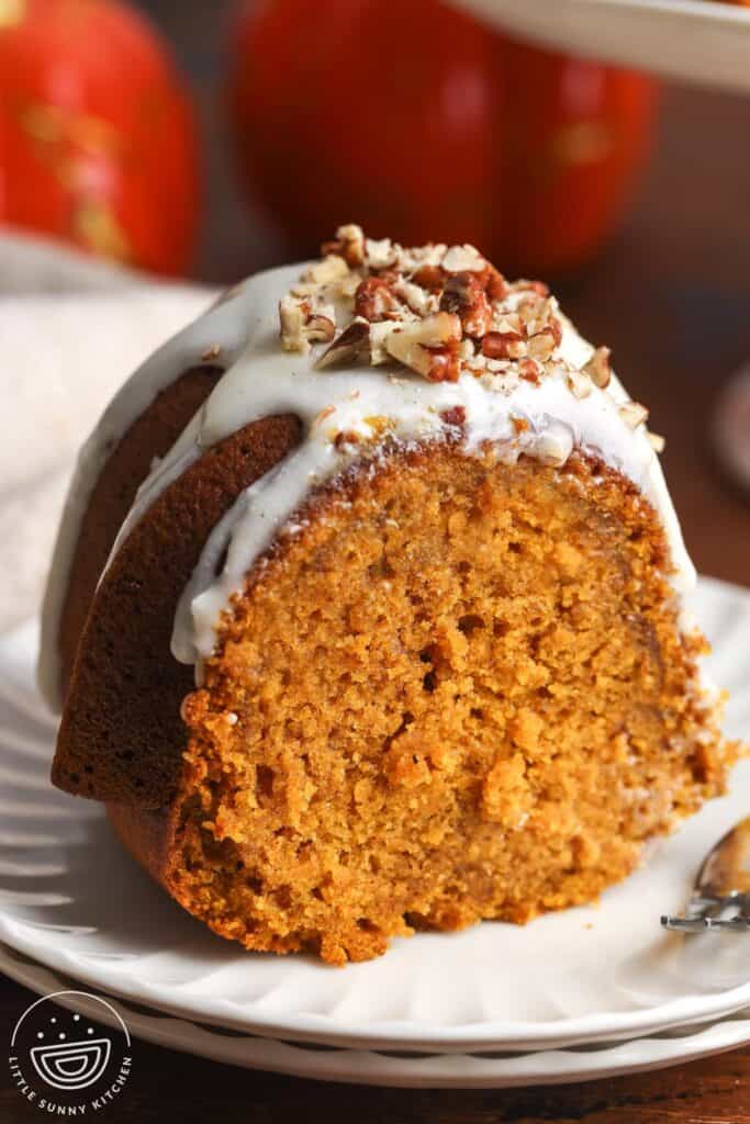 A single thick slice of pumpkin bundt cake on a white plate, topped with glaze and chopped pecans, with a fork resting beside it.