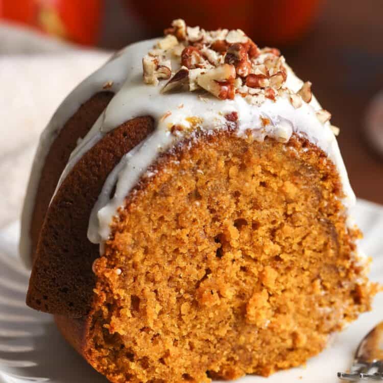 A single thick slice of pumpkin bundt cake on a white plate, topped with glaze and chopped pecans, with a fork resting beside it.