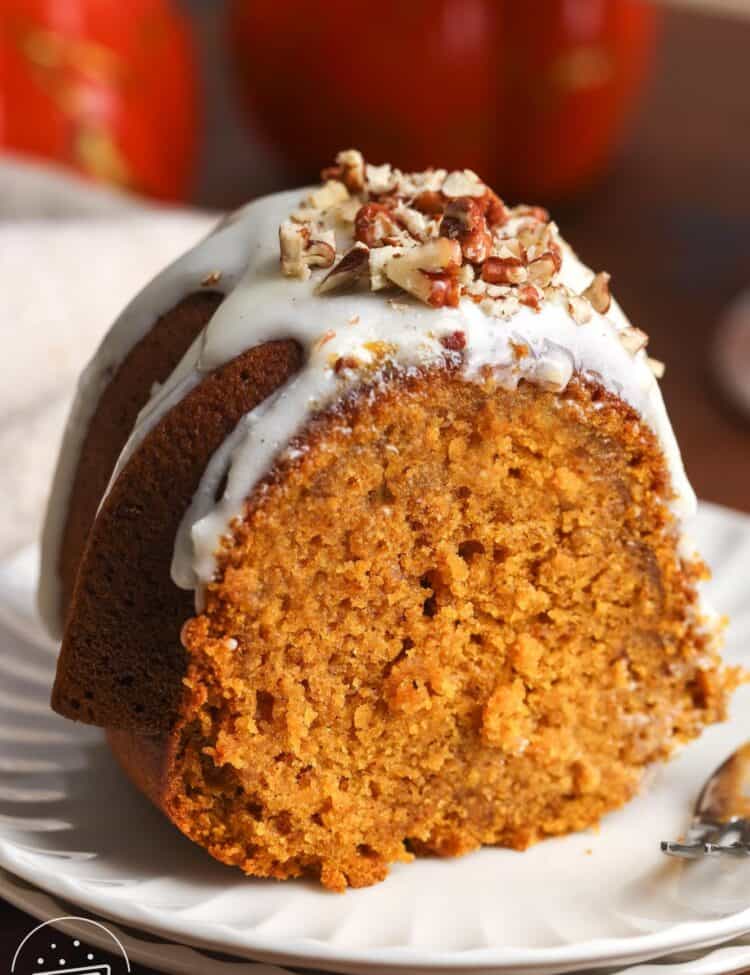 A single thick slice of pumpkin bundt cake on a white plate, topped with glaze and chopped pecans, with a fork resting beside it.