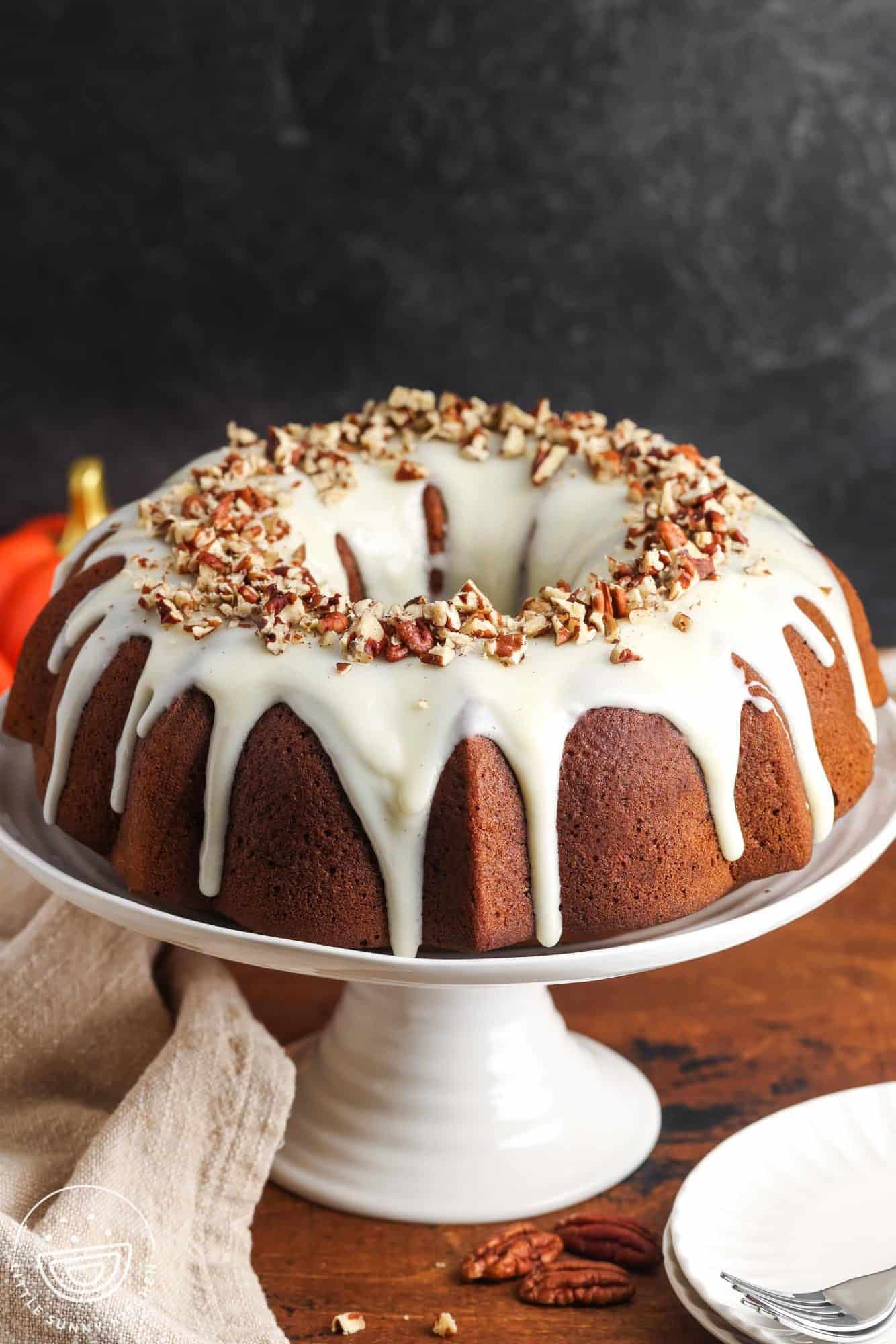 A whole pumpkin bundt cake on a white cake stand, topped with creamy white glaze dripping down the sides and a ring of chopped pecans.