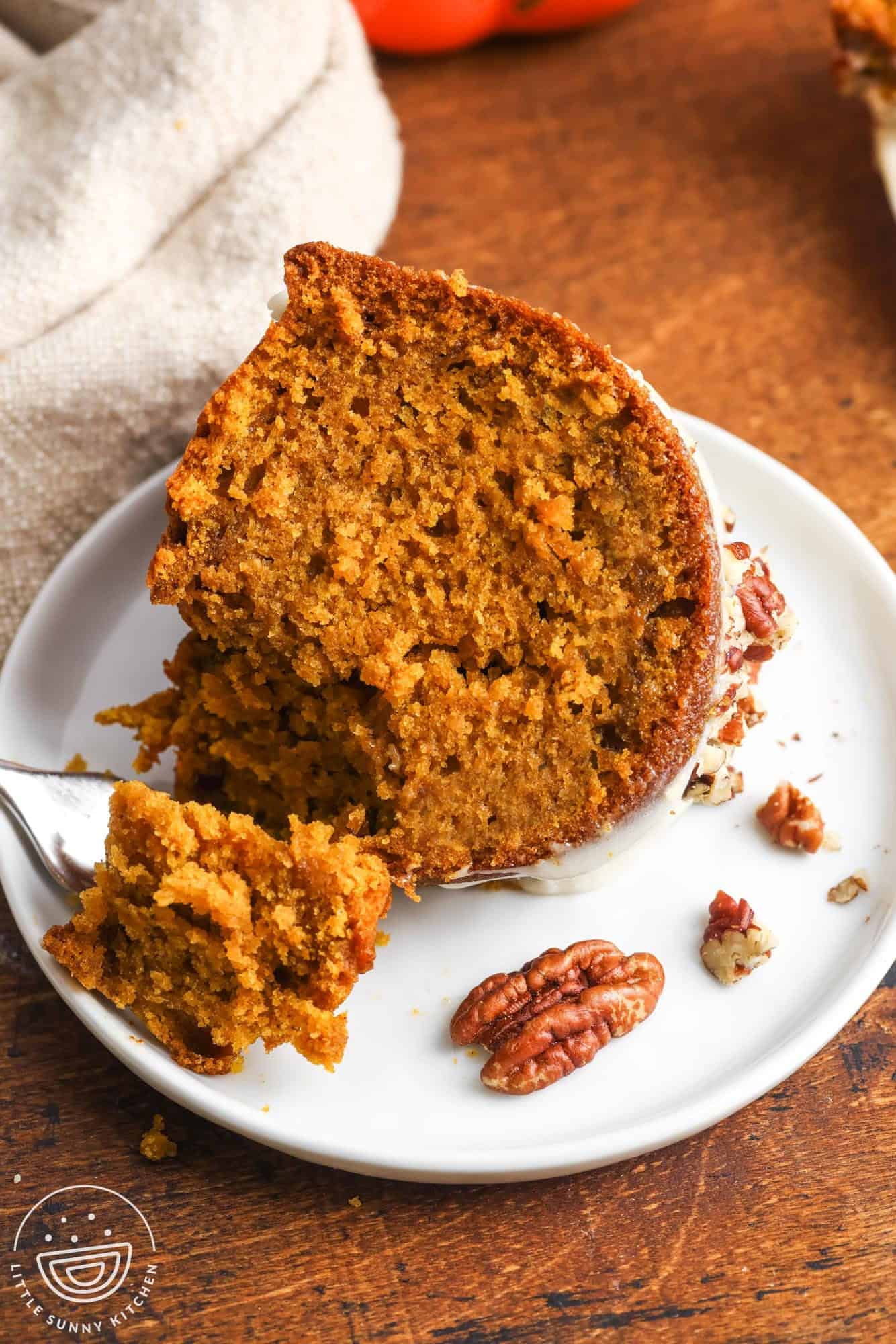 A close-up of a moist slice of pumpkin bundt cake on a white plate, with a fork breaking off a bite and pecans scattered around.