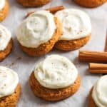 Soft pumpkin cookies topped with swirls of creamy cream cheese frosting, lightly sprinkled with cinnamon, arranged on parchment paper with cinnamon sticks scattered around.