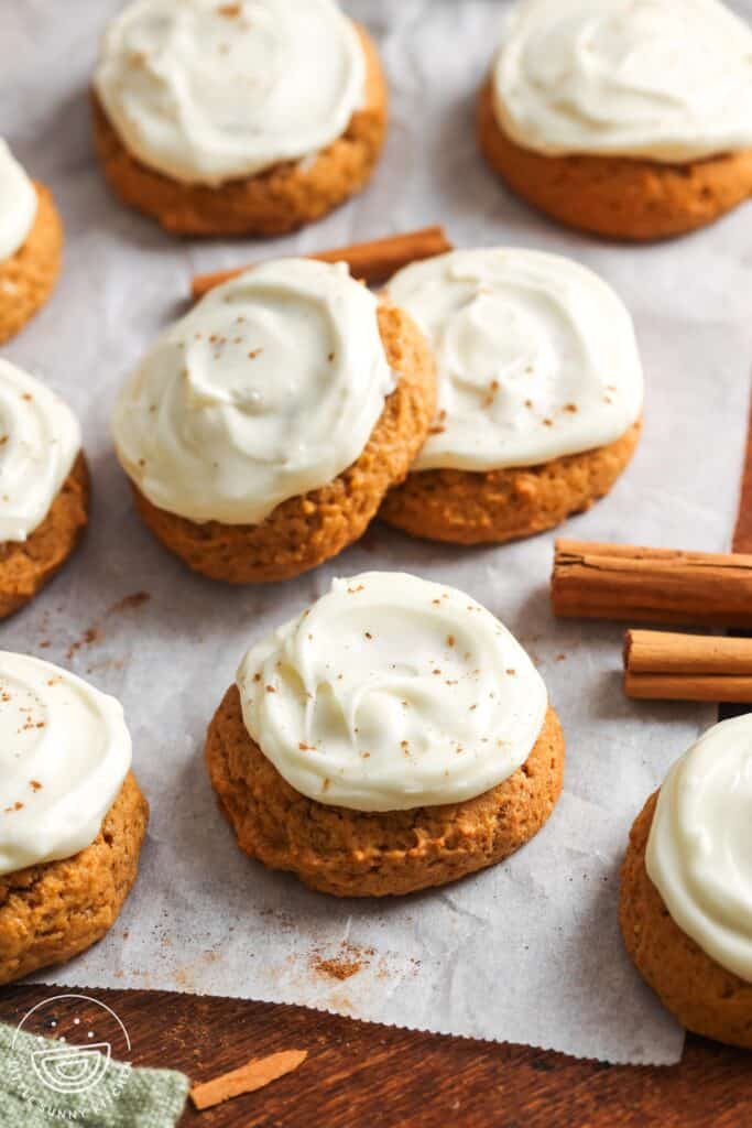 Soft pumpkin cookies topped with swirls of creamy cream cheese frosting, lightly sprinkled with cinnamon, arranged on parchment paper with cinnamon sticks scattered around.