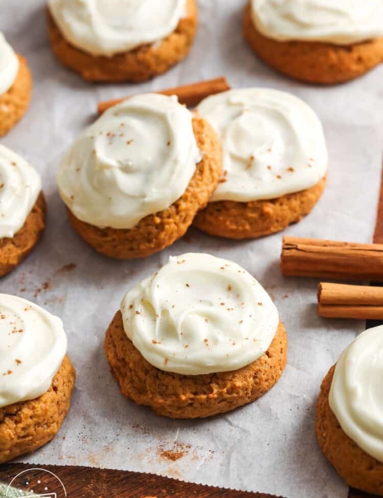 Soft pumpkin cookies topped with swirls of creamy cream cheese frosting, lightly sprinkled with cinnamon, arranged on parchment paper with cinnamon sticks scattered around.