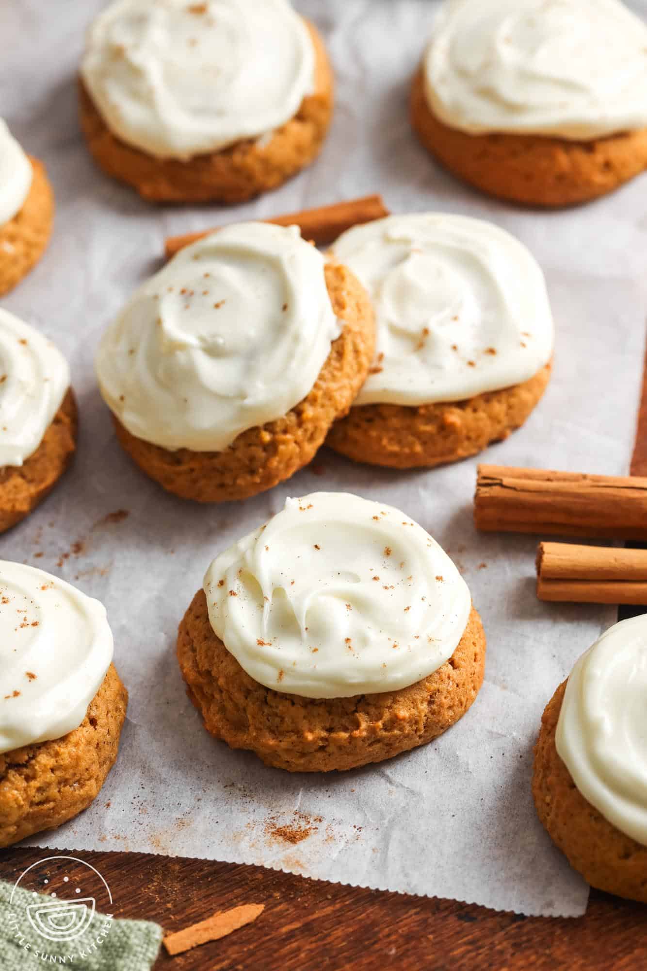 Soft pumpkin cookies topped with swirls of creamy cream cheese frosting, lightly sprinkled with cinnamon, arranged on parchment paper with cinnamon sticks scattered around.