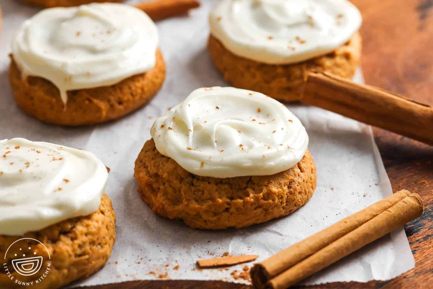 Close-up of soft pumpkin cookies topped with creamy cream cheese frosting and a light sprinkle of cinnamon, arranged on parchment paper with cinnamon sticks on the side.