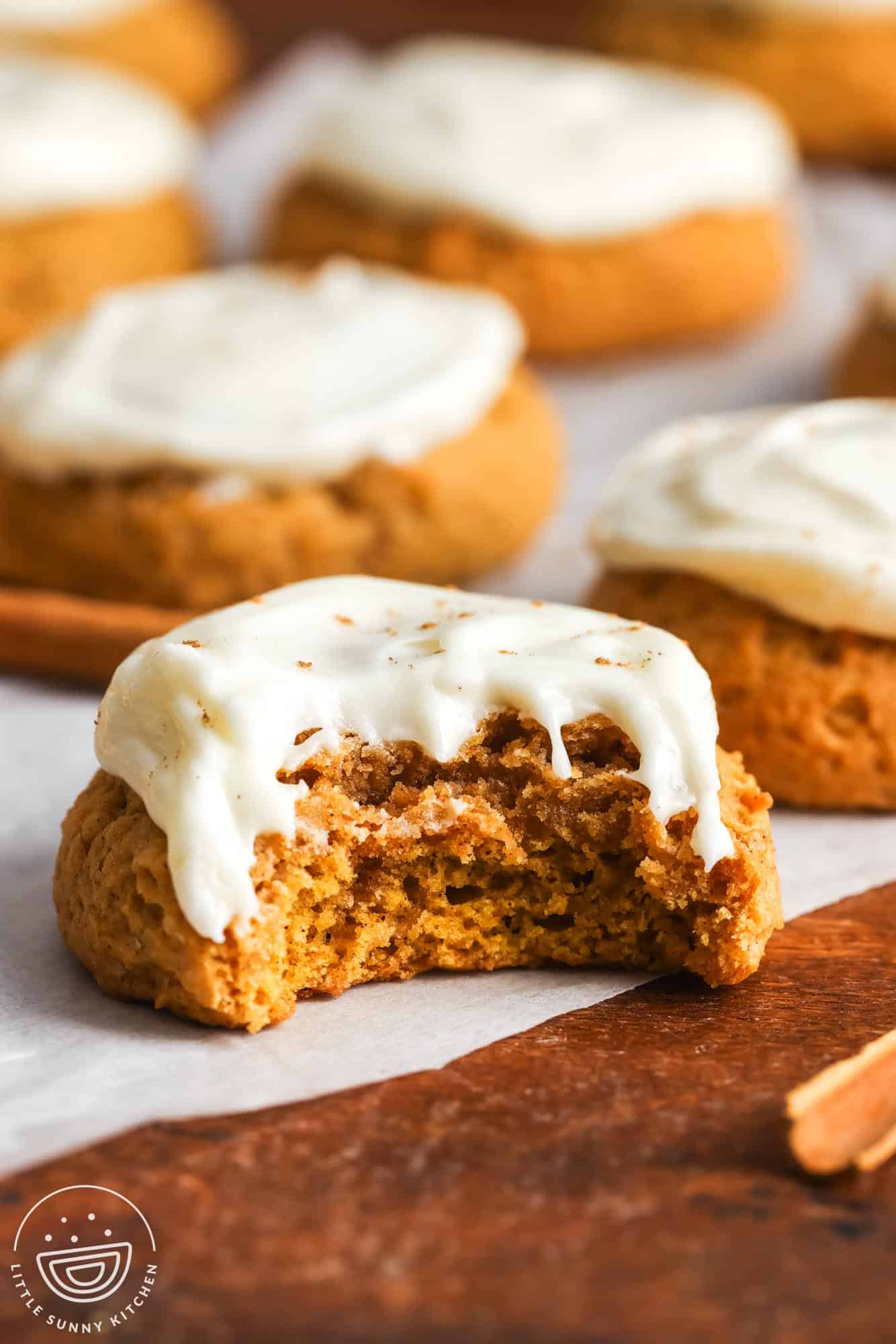 Close-up of a soft pumpkin cookie with creamy white frosting, showing a large bite taken out to reveal the moist, fluffy inside. Other frosted cookies are blurred in the background on parchment paper.