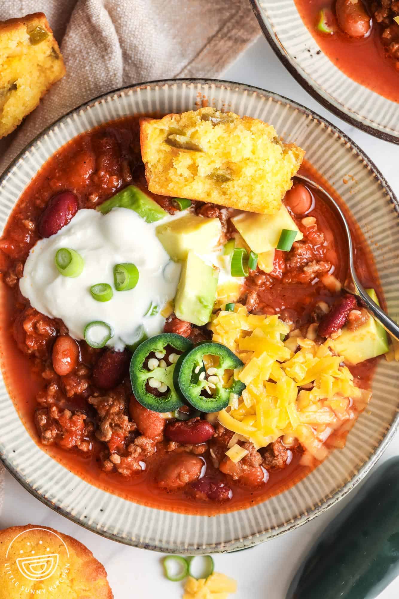 A ceramic bowl of chili, topped with cornbread, sour cream, cheese, avocado, and jalapeno.