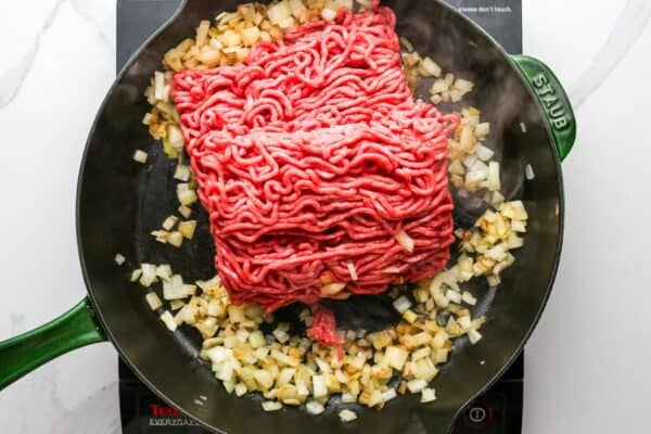 raw ground beef added to a skillet of cooked diced onions.