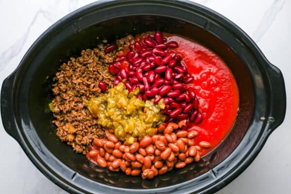 an oval crockpot filled with cooked ground beef, pinto beans, kidney beans, canned chilies and tomato sauce.