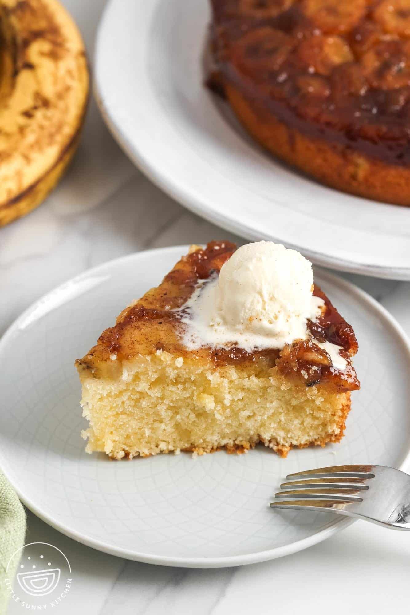 A plated slice of banana upside down cake with vanilla ice cream, with the full cake in the background.