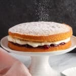 a two layer victoria sponge cake with raspberry and cream filling, on a white cake pedestal. Powdered sugar is being sifted over the top of the unfrosted cake.