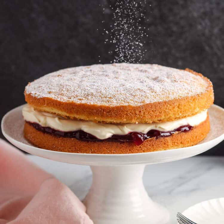 a two layer victoria sponge cake with raspberry and cream filling, on a white cake pedestal. Powdered sugar is being sifted over the top of the unfrosted cake.