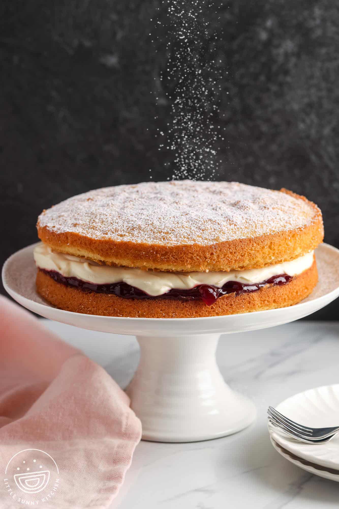 a two layer victoria sponge cake with raspberry and cream filling, on a white cake pedestal. Powdered sugar is being sifted over the top of the unfrosted cake.