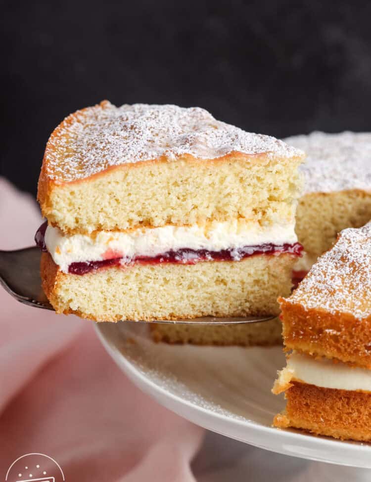 a slice of British sponge cake lifted from the rest of the cake. The side view of the slice shows the layers of whipped cream and raspberry jam filling.