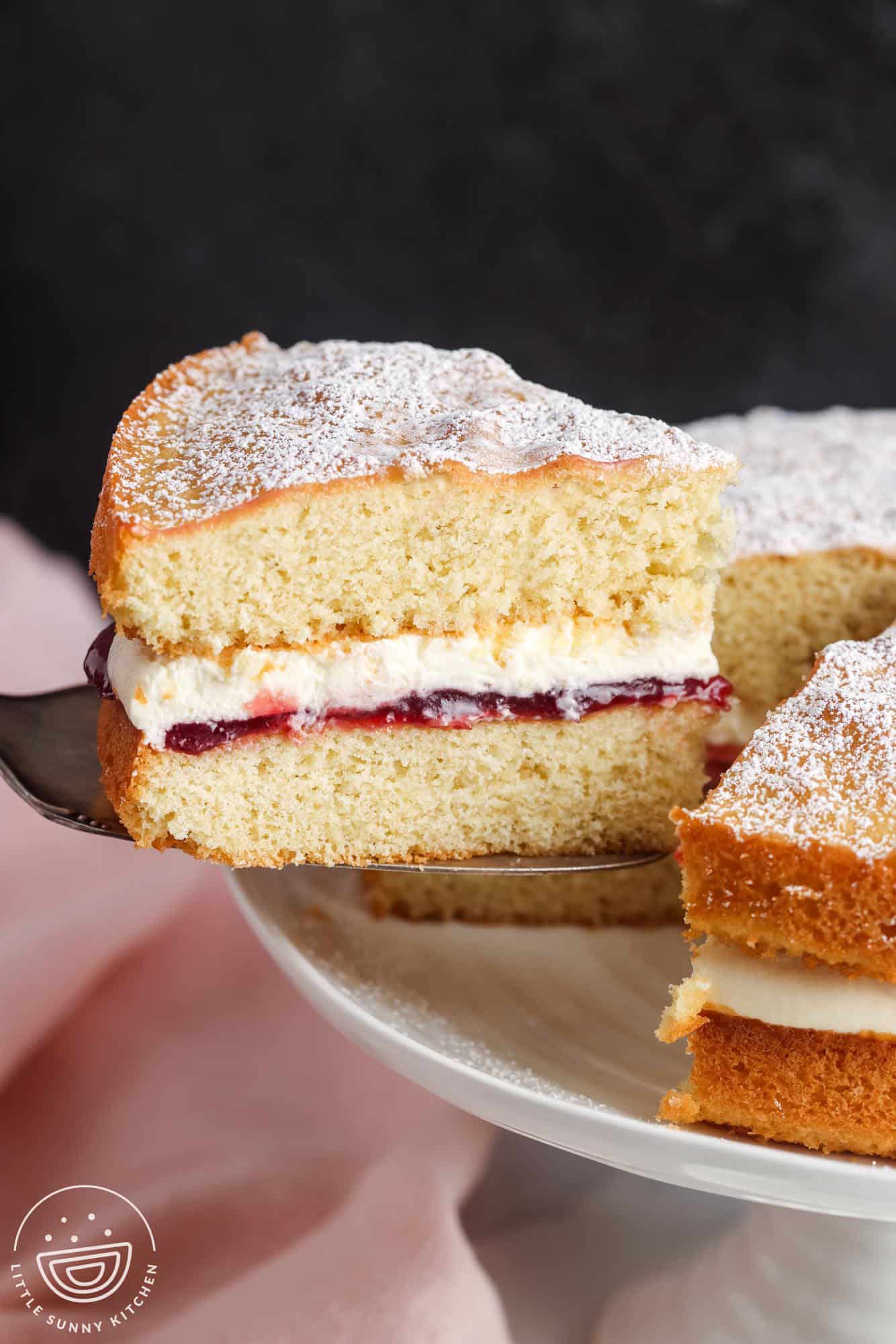 a slice of British sponge cake lifted from the rest of the cake. The side view of the slice shows the layers of whipped cream and raspberry jam filling.
