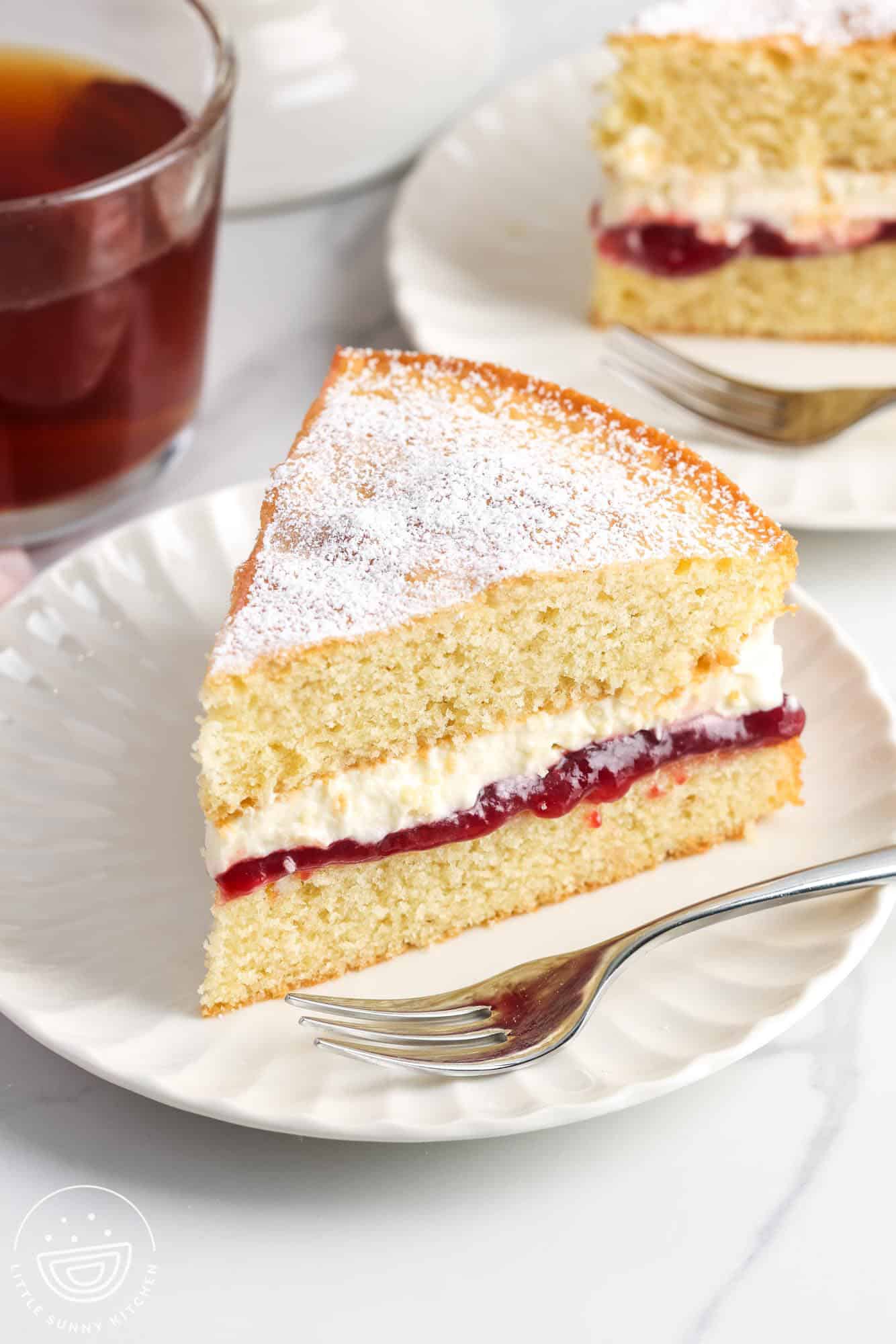 a wedge shaped slice of victoria sponge cake on a small plate with a dessert fork. it's a two layer cake with raspberry filling and whipped cream filling, dusted with powdered sugar.