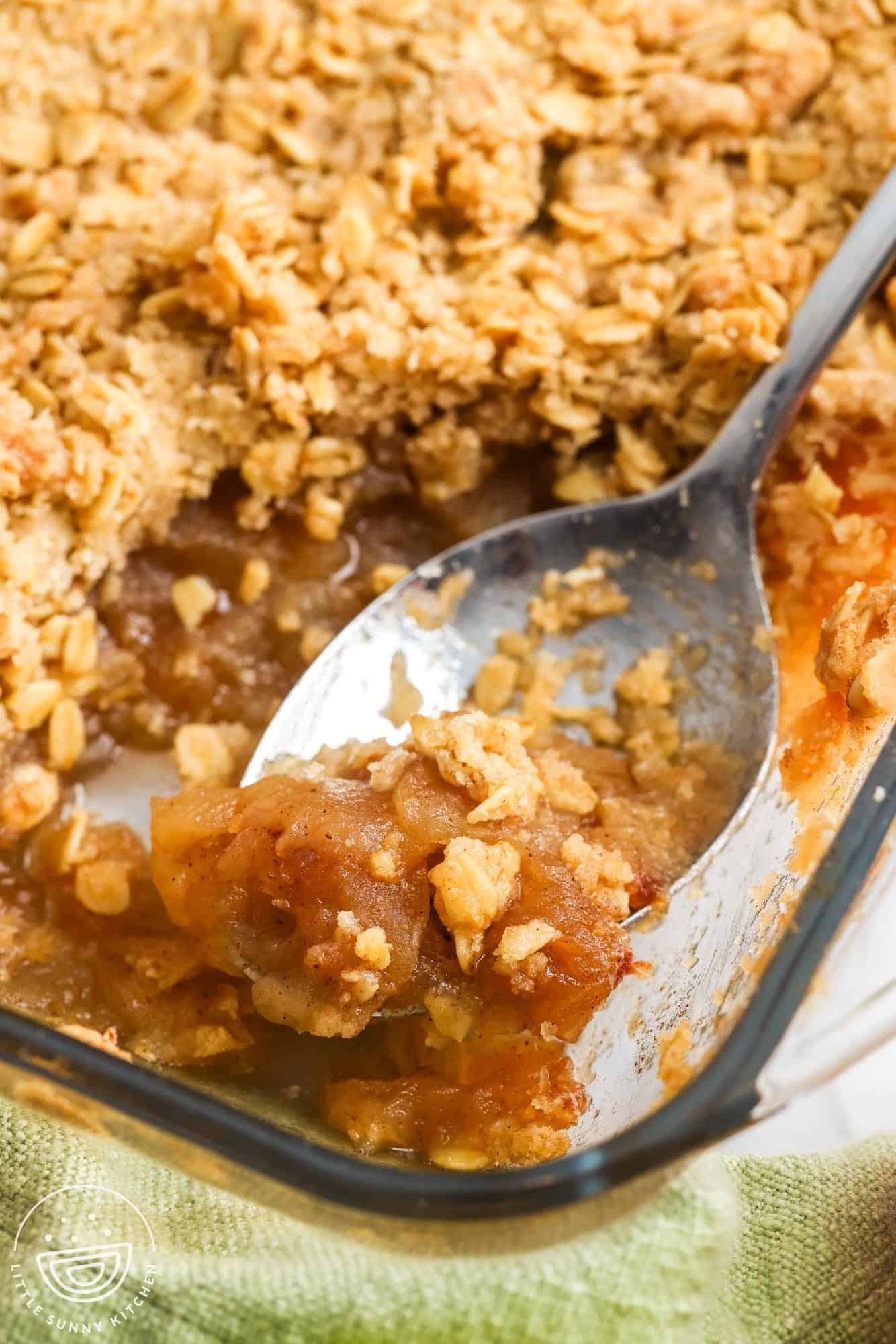 baked apple crisp with crumble oat topping in a pan. A spoon is picking up a serving to show the soft cinnamon apples underneath the topping.