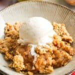 a bowl of homemade apple crisp topped with a scoop of vanilla ice cream. in the background are fresh apples.