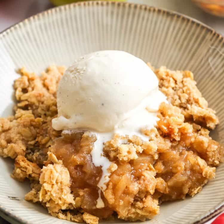 a bowl of homemade apple crisp topped with a scoop of vanilla ice cream. in the background are fresh apples.