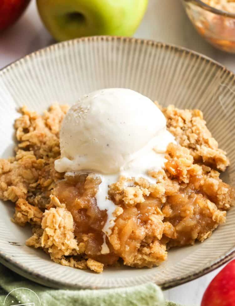 a bowl of homemade apple crisp topped with a scoop of vanilla ice cream. in the background are fresh apples.