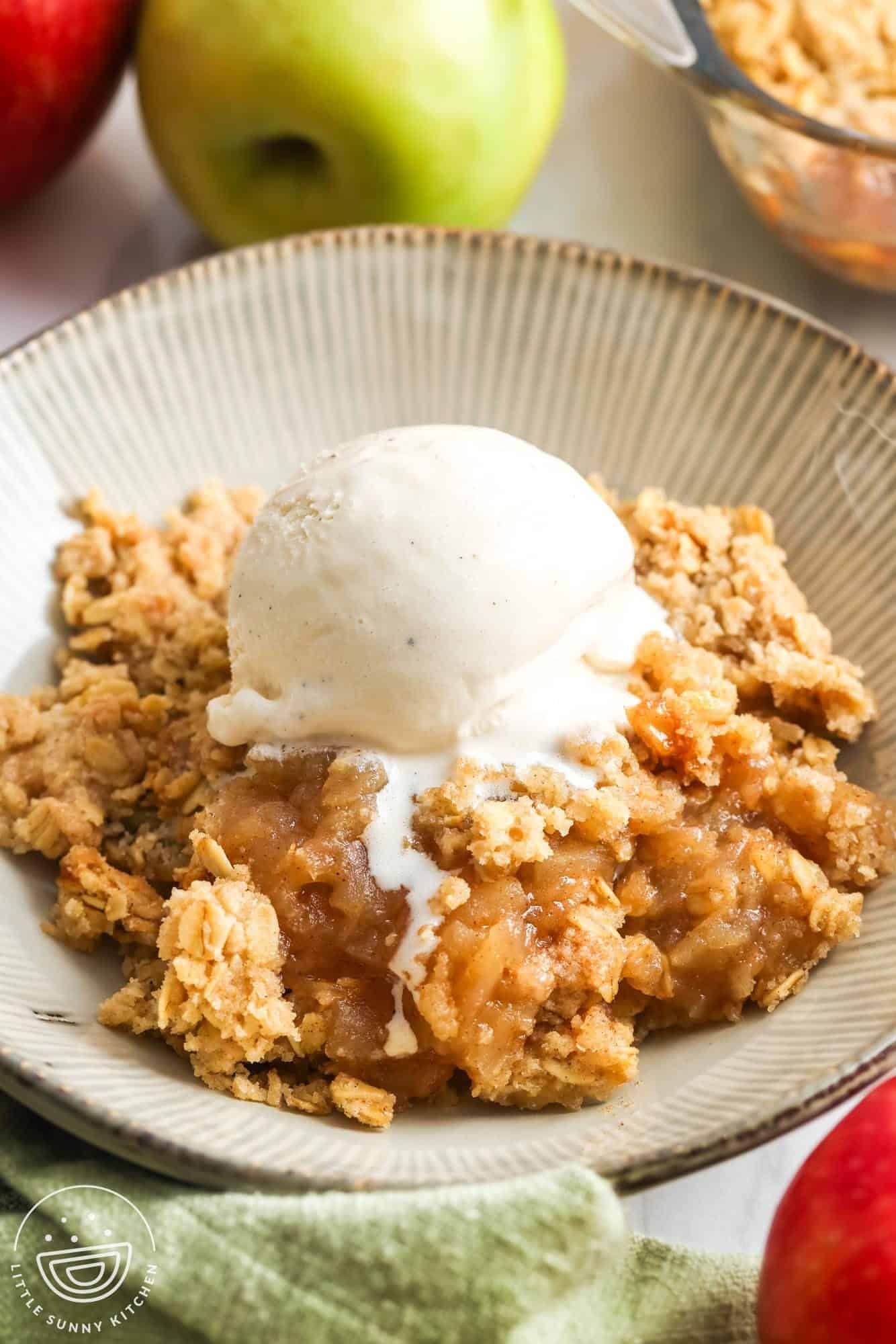 a bowl of homemade apple crisp topped with a scoop of vanilla ice cream. in the background are fresh apples.