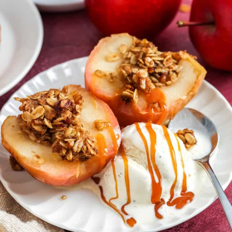 Baked apple halves filled with oat walnut crumble on a white plate, served with vanilla ice cream and caramel drizzle; red apples in the background.