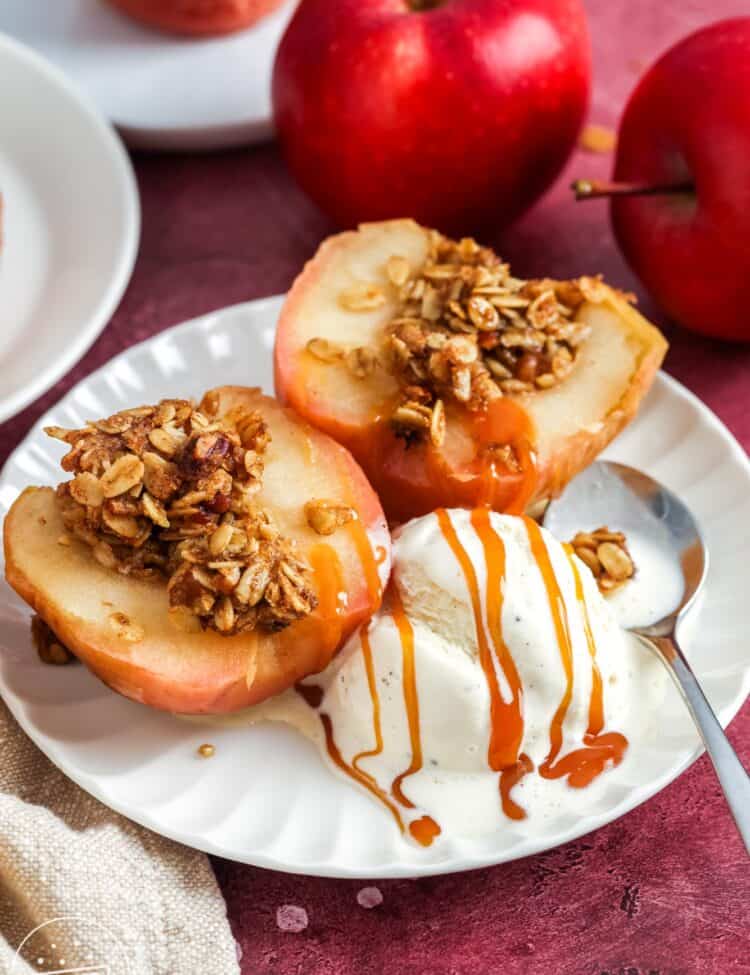 Baked apple halves filled with oat walnut crumble on a white plate, served with vanilla ice cream and caramel drizzle; red apples in the background.