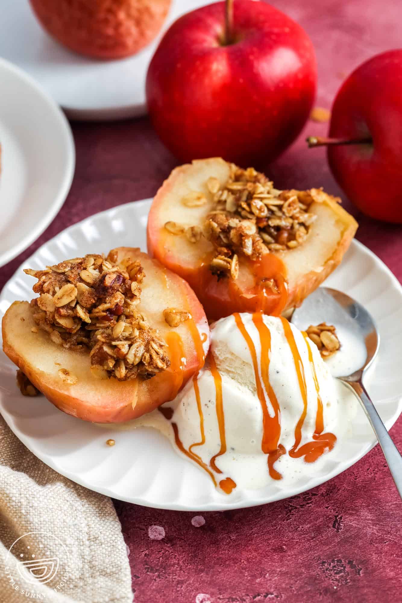 Baked apple halves filled with oat walnut crumble on a white plate, served with vanilla ice cream and caramel drizzle; red apples in the background.