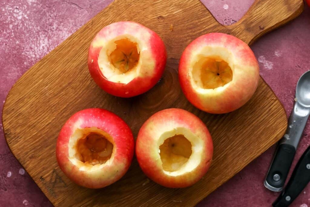Four red apples cored and hollowed on a wooden cutting board, ready for filling.
