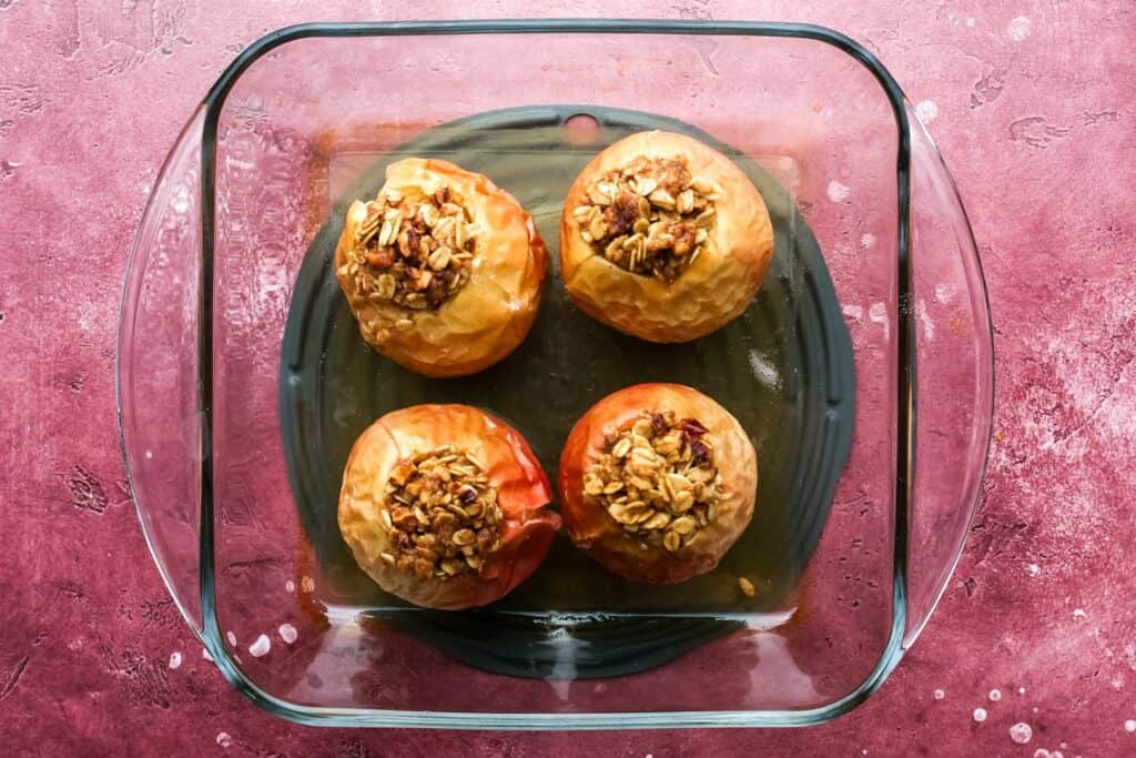 Fully baked apples with golden oat topping in a glass dish against a pink background.