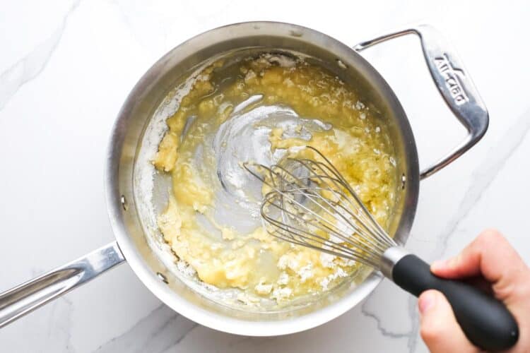 a whisk stirring a roux of butter and flour in a metal saucepan.