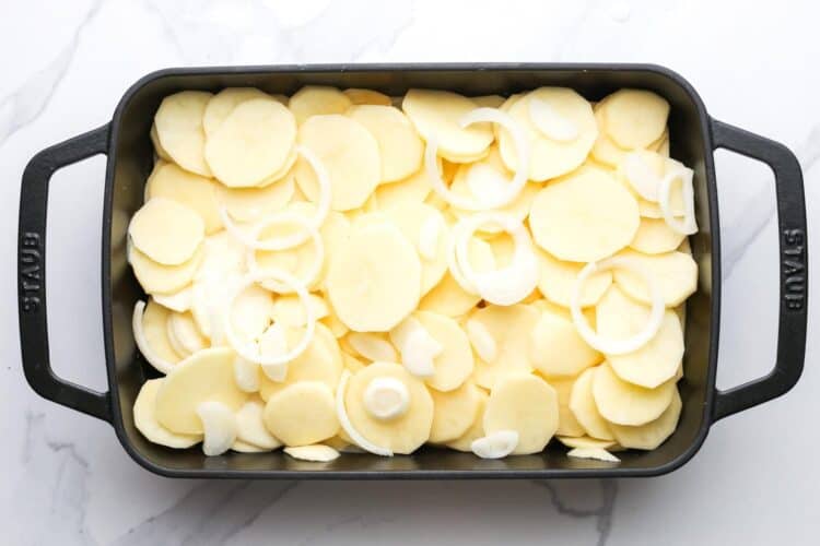 thinly sliced potatoes and onions arranged in a black rectangular baking dish.